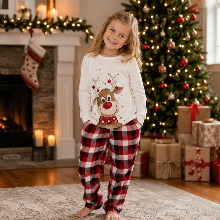 A young girl stands barefoot in a living room wearing a white long-sleeve Christmas pajama top with a reindeer graphic and red-and-black plaid pajama pants. A decorated Christmas tree, fireplace, stockings, and wrapped gifts are visible in the background.